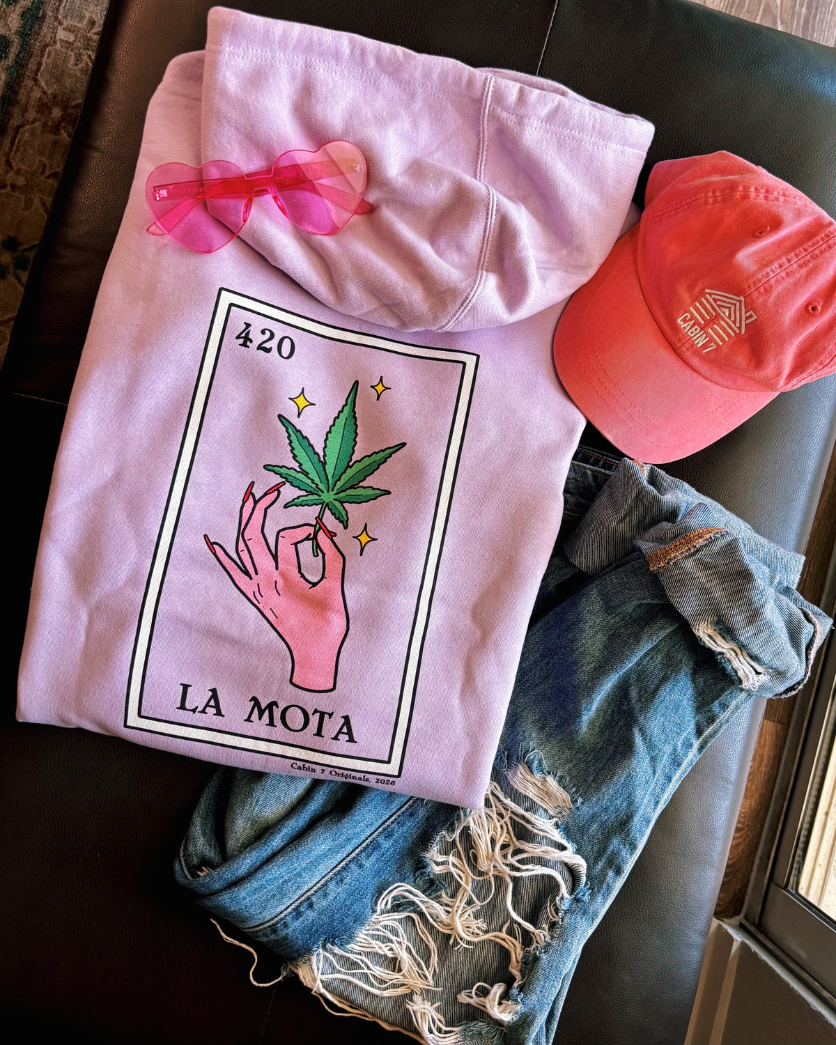 A folded light pink hoodie with a colorful graphic print of a hand holding a cannabis leaf and the text '420 LA MOTA'