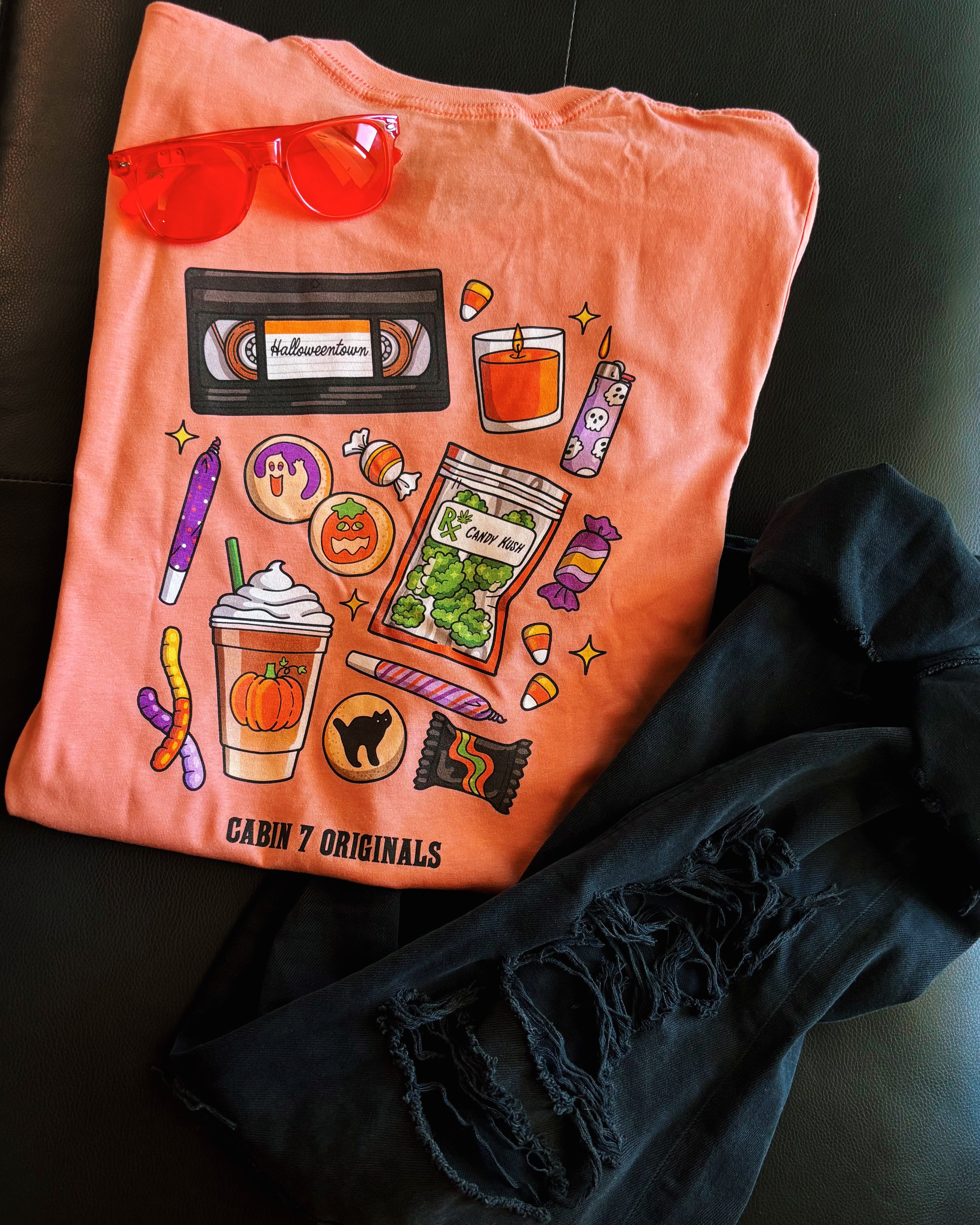 An orange t-shirt with a bright colorful stoner Halloween design, featuring several items such as a candle, a lighter, a VHS tape, and a bag of cannabis.