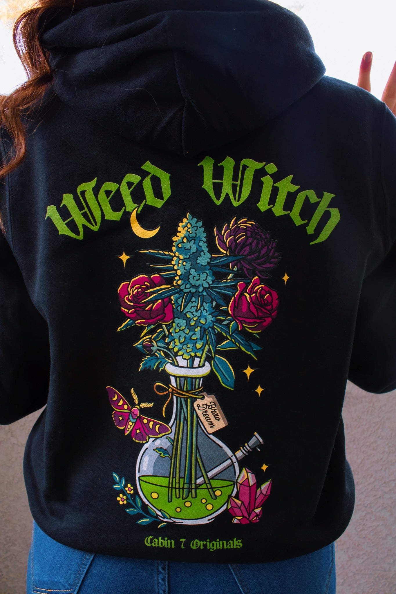 A black hoodie with a "Weed Witch" design, featuring a bong filled with flowers and a cannabis plant, as well as a moth, a moon, and crystals. Weed witch apparel, stoner Halloween