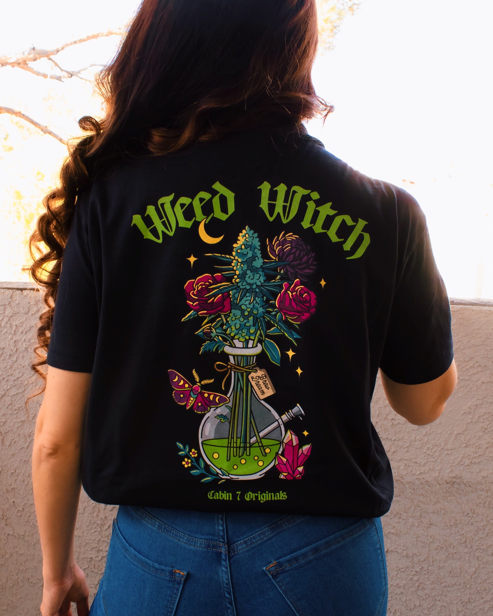 A black t-shirt with lime green text reading "Weed Witch" and a bong full of flowers and cannabis. Stoner witch apparel, spooky stoner apparel, hippie Halloween apparel.