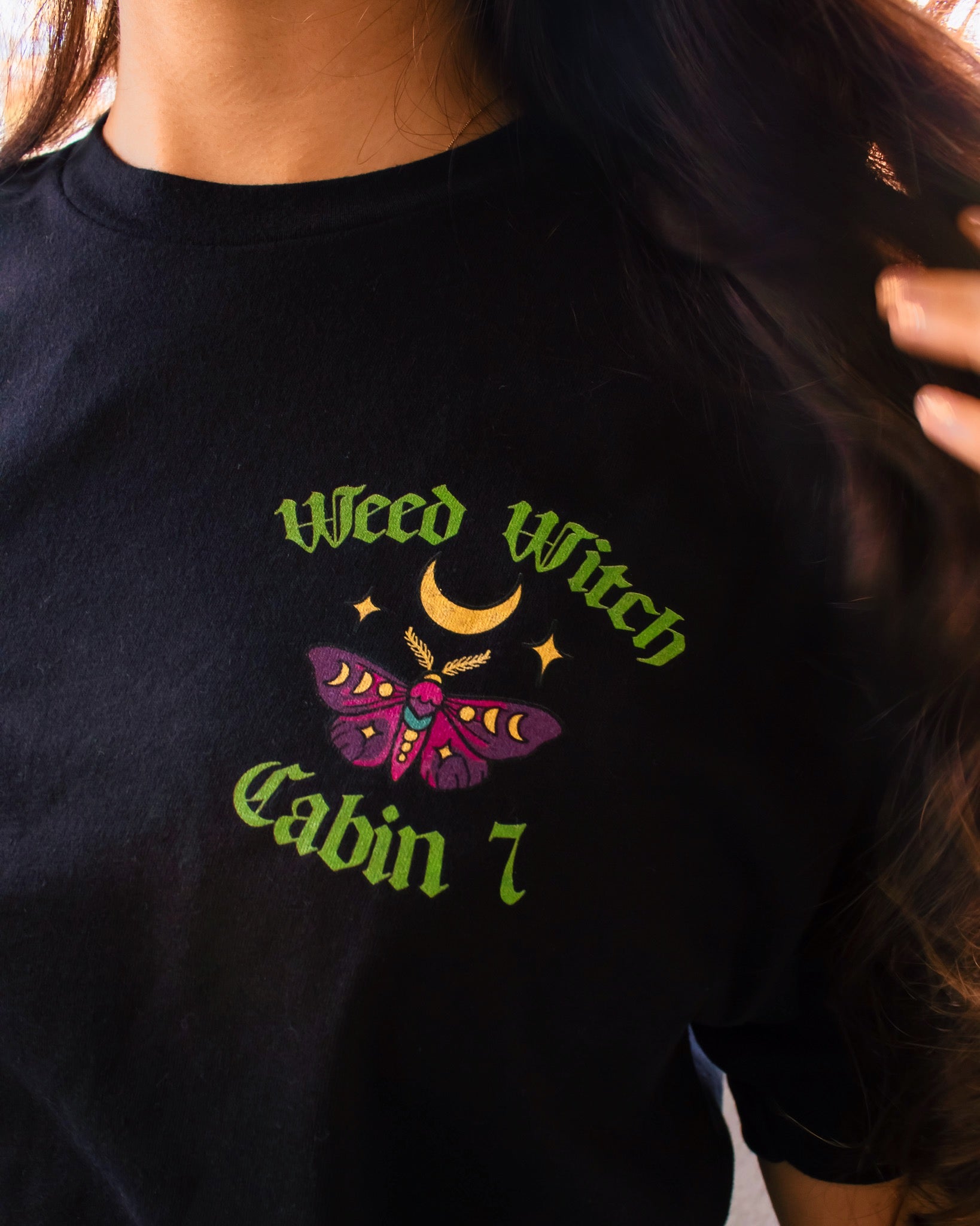 A black t-shirt with lime green text reading "Weed Witch" and a moth with a moon. Stoner witch apparel, spooky stoner apparel, hippie Halloween apparel.