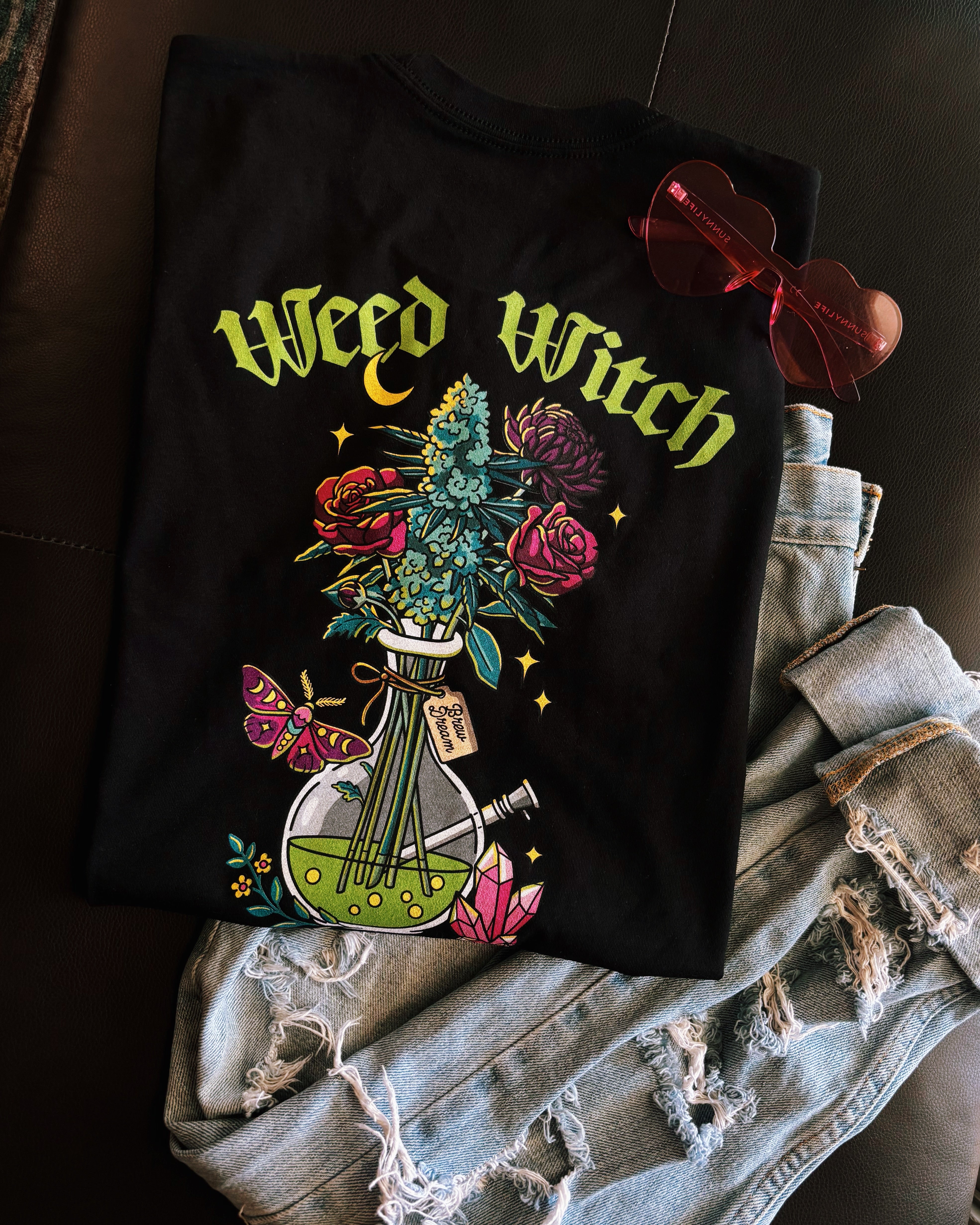 A black t-shirt with lime green text reading "Weed Witch" and a bong full of flowers and cannabis. Stoner witch apparel, spooky stoner apparel, hippie Halloween apparel.