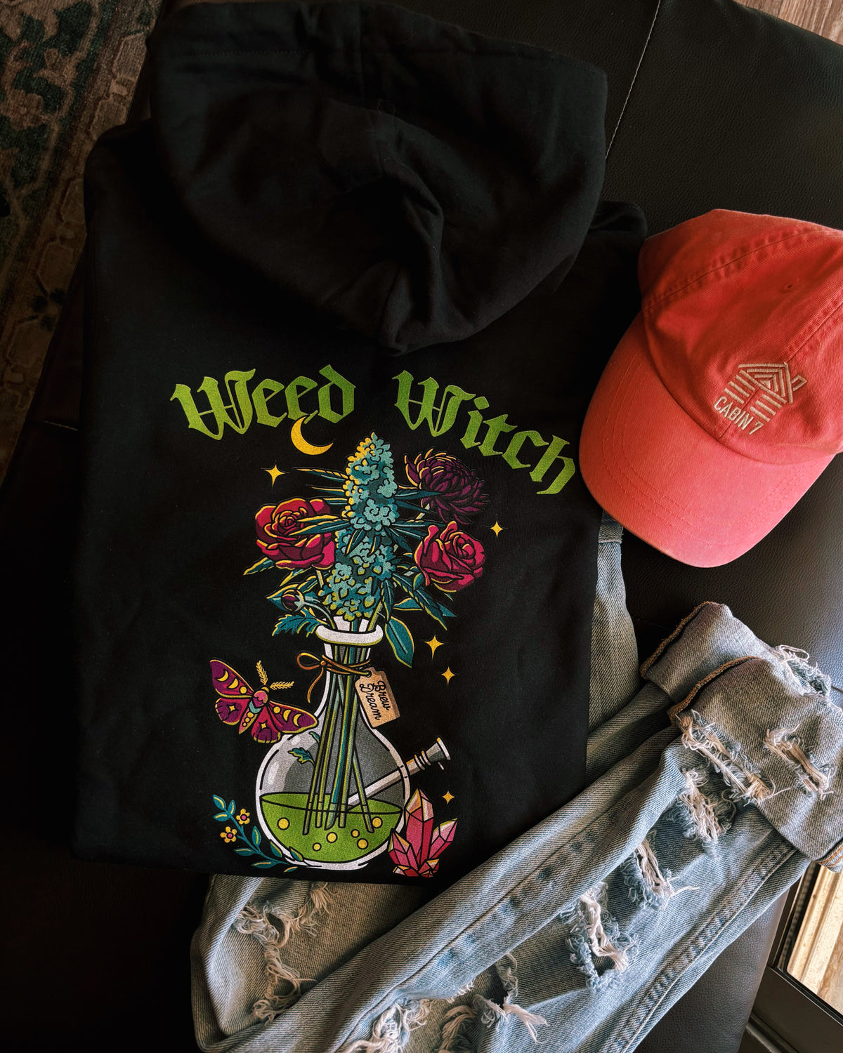 A black hoodie with a "Weed Witch" design, featuring a bong filled with flowers and a cannabis plant, as well as a moth, a moon, and crystals. Weed witch apparel, stoner Halloween