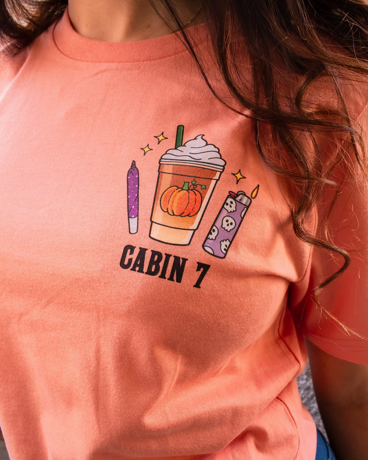 An orange t-shirt with a bright colorful stoner design, featuring several items such as a candle, a lighter, and a pumpkin spice latte. 