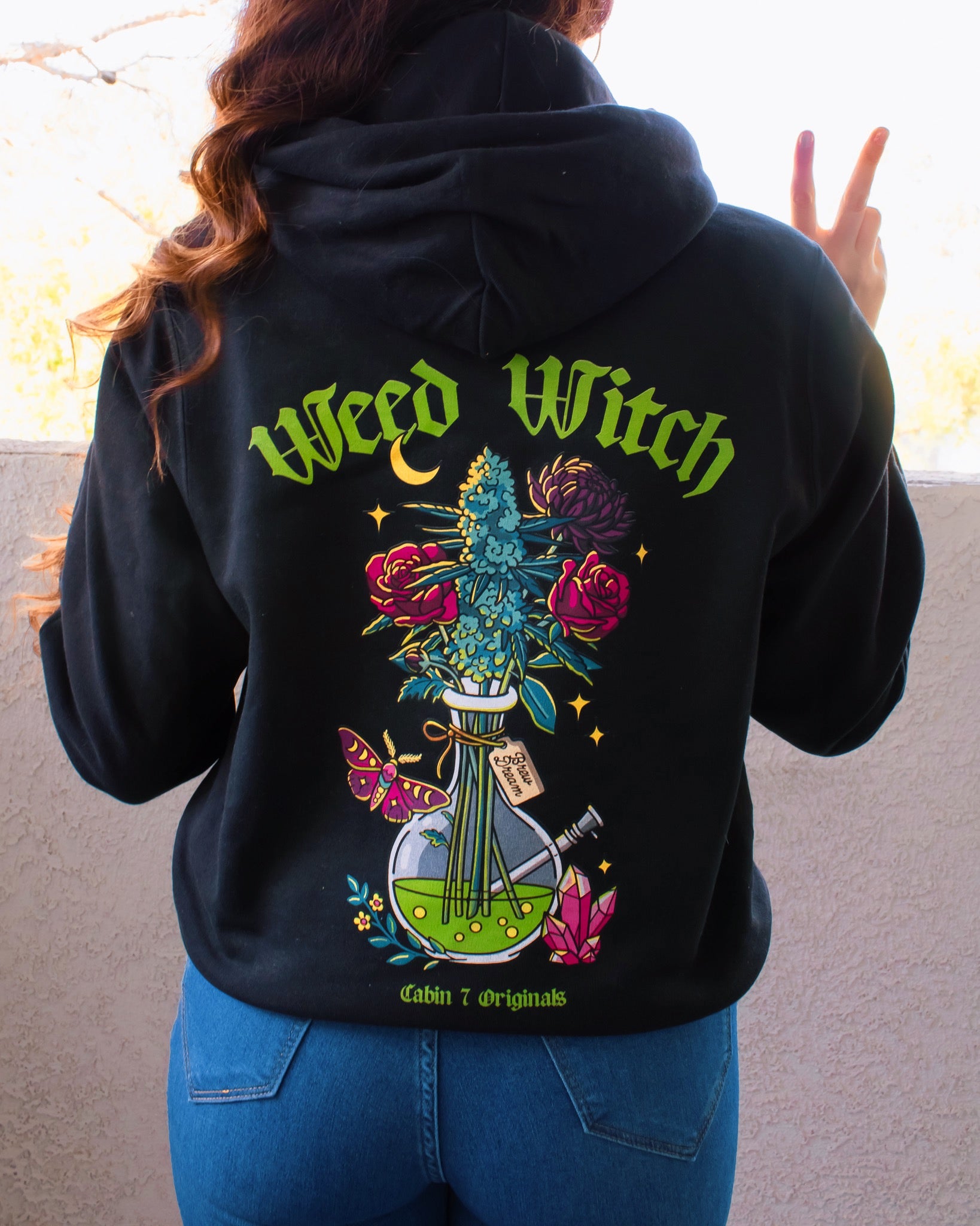 A black hoodie with a "Weed Witch" design, featuring a bong filled with flowers and a cannabis plant, as well as a moth, a moon, and crystals. Weed witch apparel, stoner Halloween