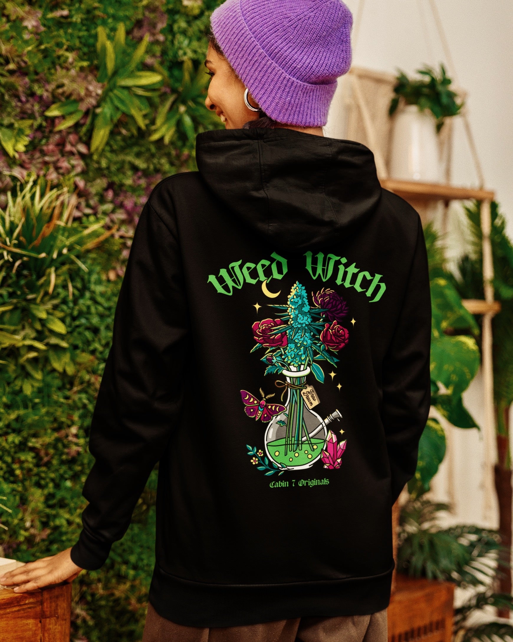 A black hoodie with a "Weed Witch" design, featuring a bong filled with flowers and a cannabis plant, as well as a moth, a moon, and crystals. Weed witch apparel, stoner Halloween