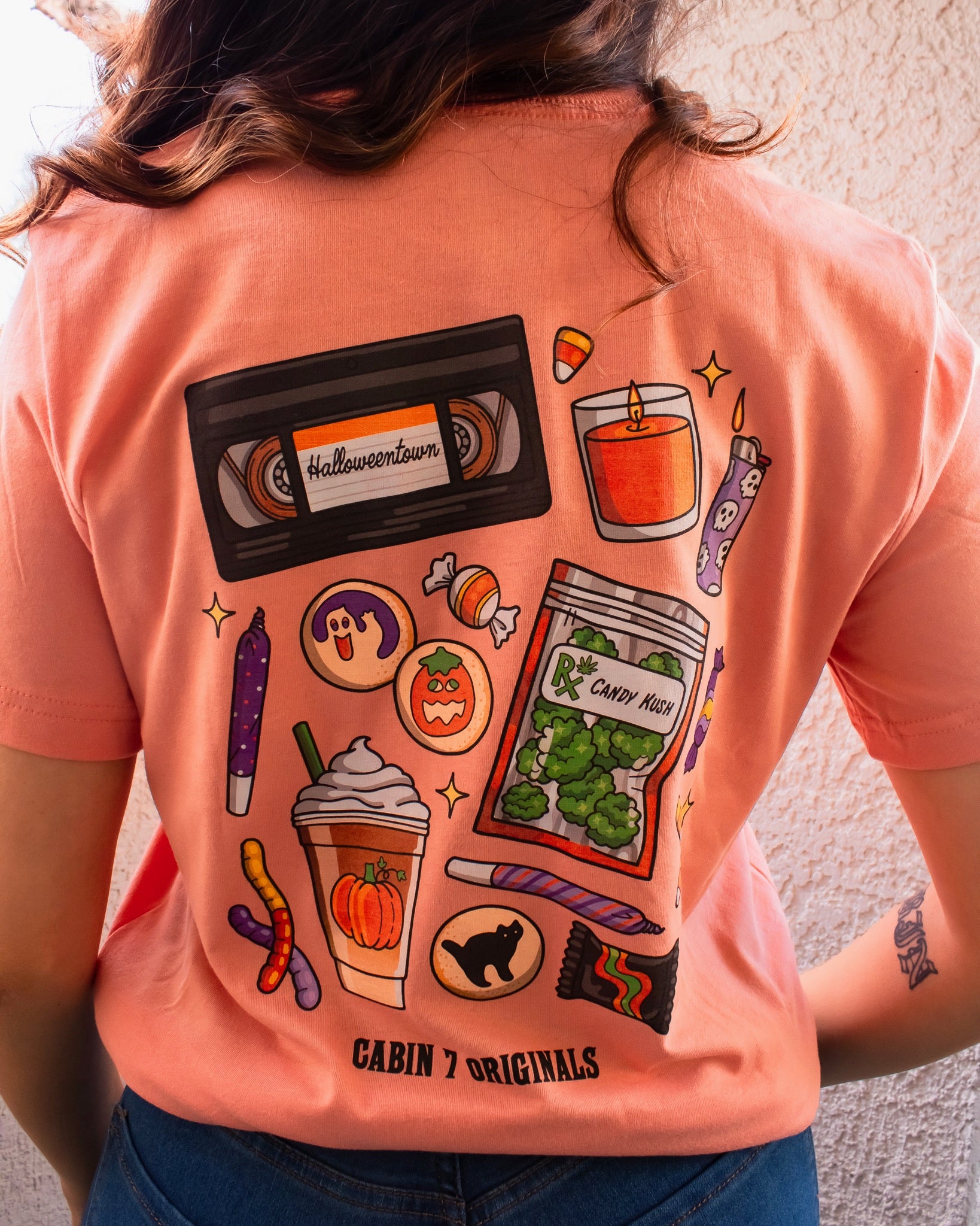 An orange t-shirt with a bright colorful stoner Halloween design, featuring several items such as a candle, a lighter, a VHS tape, and a bag of cannabis. 